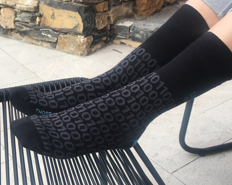 Op de afbeelding: Een paar zwarte sokken met een herhalend patroon van grijze 1s en 0s. De sokken liggen op een zwarte stoel.