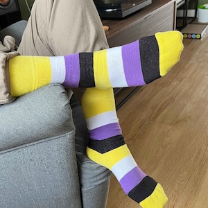 Puede incluir: Un par de calcetines con rayas amarillas, blancas, moradas y negras. Los calcetines están puestos en los pies de una persona.