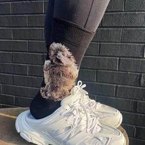 May include: White Balenciaga trainers with a chunky sole and lace-up fastening. The trainers are worn with black socks featuring a brown and white animal design. The word "Balenciaga" is printed on the side of the shoe.