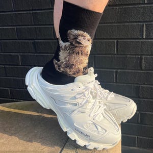 May include: White Balenciaga trainers with a thick sole and laces. The trainers are worn with black socks featuring a detailed illustration of a bird. The word "Balenciaga" is printed on the side of the shoe.