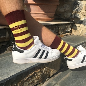 Könnte beinhalten: Ein Paar weiße Sneaker mit schwarzen Streifen und dem Schriftzug "SUPERSTAR" an der Seite. Die Person trägt rotbraune und gelbe gestreifte Socken mit dem Text "JAMES is a free elf" auf einem Socken.