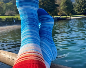 Könnte beinhalten: Ein Paar blau-weiß gestreifte Socken mit rotem Bund. Die Socken liegen auf einem Holzgeländer mit einem See und Bäumen im Hintergrund.