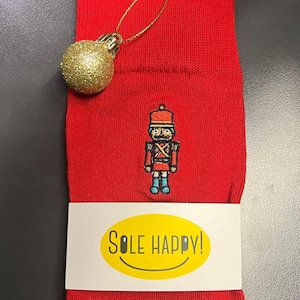May include: Bright red socks featuring a nutcracker motif and the text "Sole Happy!" on a white label. A gold glitter bauble hangs from the top. A festive Christmas item.