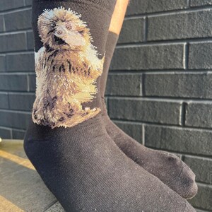 May include: A pair of black crew socks featuring a detailed, woven graphic of a fluffy, light brown dog standing on its hind legs. The socks have a ribbed cuff at the top and a solid black foot section, showcasing a clear, textured animal portrait.