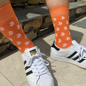 Puede incluir: Un par de zapatillas blancas con rayas negras y calcetines naranjas con un patrón de símbolo Bitcoin blanco.