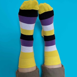 Peut inclure: Une paire de chaussettes rayées jaunes, noires, violettes et blanches. Les chaussettes sont pliées en haut et sur un fond bleu.