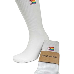 Op de afbeelding: Een paar witte sokken met een klein, kleurrijk regenboogvlaggetje geborduurd aan de zijkant. De sokken zijn omlaag gerold en de verpakking zegt "seriously good socks".