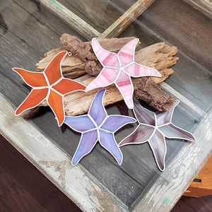 May include: Four stained glass starfish ornaments in different colours: orange, pink, purple, and brown. The ornaments are displayed on a piece of driftwood.
