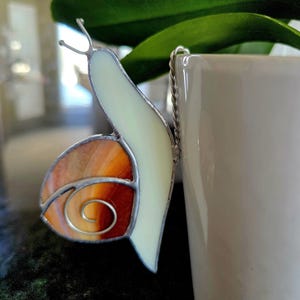 May include: A stained glass snail ornament with a cream-coloured body and an orange and brown shell. The snail has a silver wire outline and a silver chain for hanging. The ornament is placed next to a white ceramic pot.