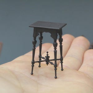 May include: A miniature black table with a square top and ornate, turned legs. The table features decorative elements and a lower support structure. The small table is held in a hand, suggesting a dollhouse or miniature setting.
