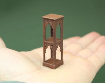 May include: A miniature brown decorative stand with an ornate, gothic-style design. The stand has a square top and base, with two shelves and arched openings. The stand is held in a hand.