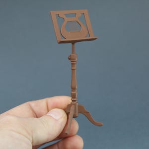 May include: A miniature brown music stand with a lyre-shaped cutout on the top. The stand has a three-legged base and a decorative, turned column. The stand is held in a person's hand against a solid blue-gray background.
