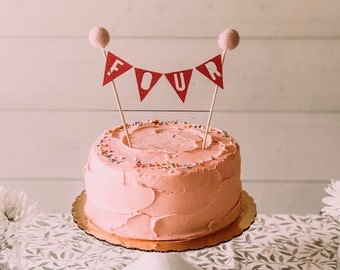 May include: A pink frosted cake with colorful sprinkles on top. The cake is decorated with a red and white banner that says "FOUR".