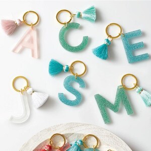 May include: A collection of letter keychains made from glittery resin in shades of pink, teal, and clear, each attached to a gold-toned metal ring with a matching fabric tassel. The letters include A, C, E, J, S, and M, with additional letters visible.