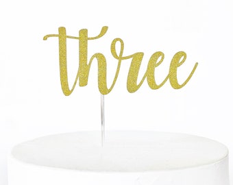 May include: Gold glitter cake topper with the word "three" in a cursive font.