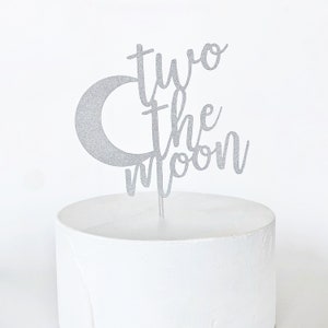 May include: Silver glitter cake topper with a crescent moon and the text "two the moon".