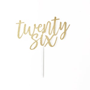 Könnte beinhalten: Goldener Glitzer-Kuchentopper mit den Worten "twenty six" in einer kursivschrift.