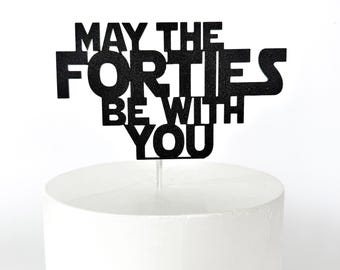 以下が含まれることがあります： 大胆なフォントで「MAY THE FORTIES BE WITH YOU」と書かれた黒いケーキトッパー。白いケーキの上にあり、40歳の誕生日のお祝いに最適です。