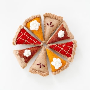May include: A circular arrangement of eight felt pie slices in various colors, including red, orange, and brown. Each slice has a crust and is decorated with embroidered details or a white flower. The pie slices are arranged on a white background.
