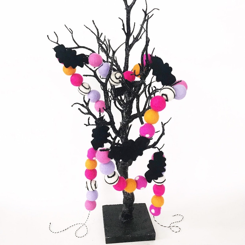 Puede incluir: Un &aacute;rbol negro brillante decorado con guirnaldas de bolas de fieltro en rosa, naranja, lavanda y blanco, y recortes de murci&eacute;lagos negros. El &aacute;rbol est&aacute; sobre una base cuadrada negra, ideal para la decoraci&oacute;n de Halloween.