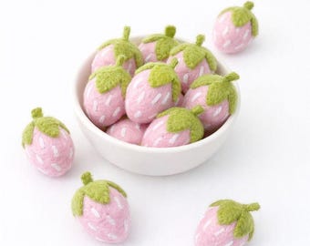 Puede incluir: Un conjunto de fresas rosas de fieltro con tapas verdes, algunas en un cuenco blanco y otras dispersas. Las fresas tienen detalles de semillas blancas sobre un fondo blanco.