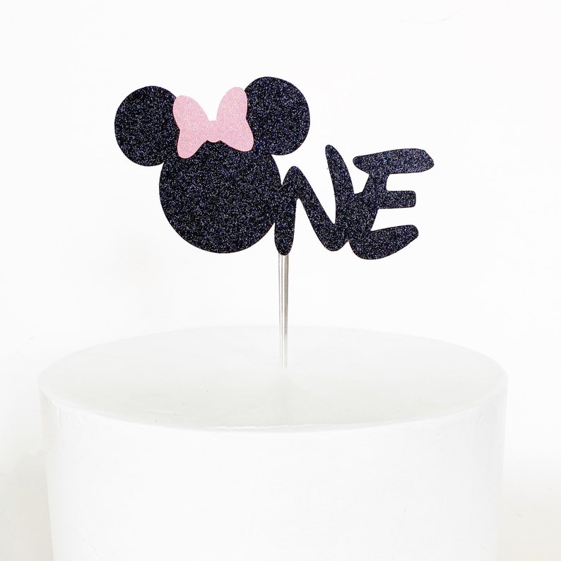 May include: Black glitter Minnie Mouse cake topper with a pink bow and the word "ONE" in black glitter.
