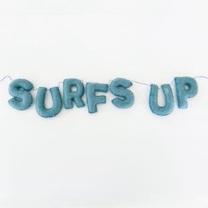 Peut inclure: Une guirlande en feutre bleu avec l'inscription "SURFS UP" cousue dans une police fantaisiste. Les lettres sont reliées par une fine ficelle noire, créant un élément décoratif. La guirlande est posée sur un fond blanc.