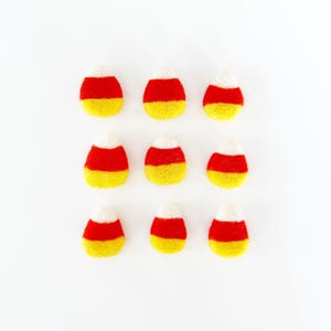 May include: Nine handmade candy corn decorations. Each piece features a white tip, a red center, and a yellow base. The candies are arranged in a grid pattern on a white background. Perfect for Halloween or fall decor.