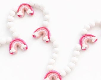 May include: A garland featuring white felt balls and pink felt rainbows with white cloud accents. The rainbows have layers of pink and peach colors. The garland is on a white background.