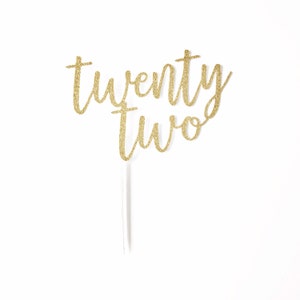 Könnte beinhalten: Goldener Glitzer-Kuchentopper mit den Worten "twenty two" in einer kursivschrift.