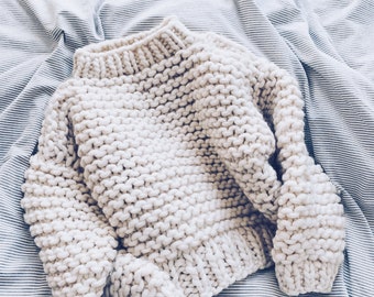 May include: A cream-colored, chunky knit sweater with a high neckline. The sweater is made of thick yarn and has a textured appearance. The sleeves are long and the garment is laying on a striped fabric.