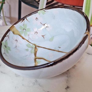 May include: A small, white ceramic bowl with a floral design and gold accents. The bowl is sitting on a white marble surface.