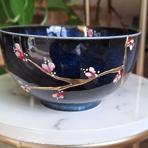 Könnte beinhalten: Eine dunkelblaue Keramikschüssel mit goldfarbener Craquelé-Glasur und einem bemalten Design eines Kirschblütenzweigs mit rosa Blüten.