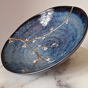 Puede incluir: Un cuenco grande de cerámica azul oscuro con un diseño floral dorado y blanco. El cuenco tiene un esmalte texturizado, moteado y un borde ligeramente irregular.