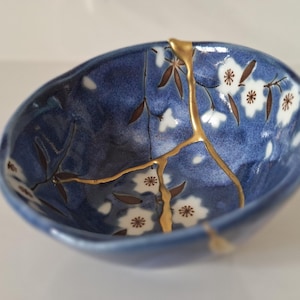 May include: A small, round ceramic bowl featuring a deep blue glaze with a white floral pattern and dark brown stems. The bowl displays a prominent crack repaired with gold lacquer, highlighting the kintsugi technique across the interior surface.