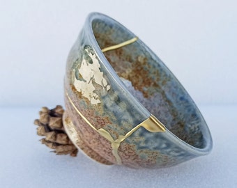 May include: A blue and brown ceramic bowl with gold accents. The bowl is cracked and repaired with gold. The bowl is sitting on a pine cone.