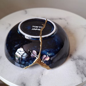 Peut inclure: Un petit bol en c&eacute;ramique bleu fonc&eacute; avec une fissure remplie de peinture dor&eacute;e. Le bol a un bord blanc et un petit motif floral peint sur le c&ocirc;t&eacute;. Le bol est marqu&eacute; "Tokyo Japan" au fond.