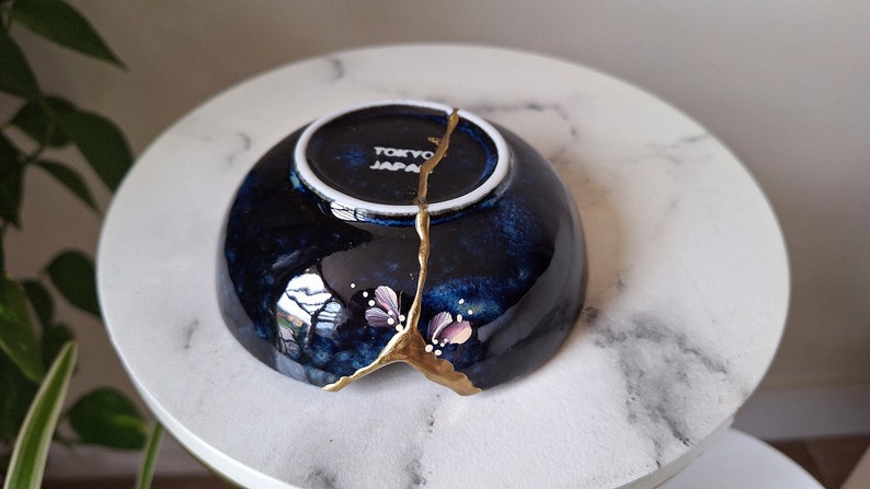 Peut inclure: Un petit bol en c&eacute;ramique bleu fonc&eacute; avec une fissure remplie de peinture dor&eacute;e. Le bol a un bord blanc et un petit motif floral peint sur le c&ocirc;t&eacute;. Le bol est marqu&eacute; "Tokyo Japan" au fond.