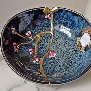 Puede incluir: Un cuenco de cerámica azul con un diseño floral dorado y rosa. El cuenco tiene un esmalte moteado y un borde dorado.