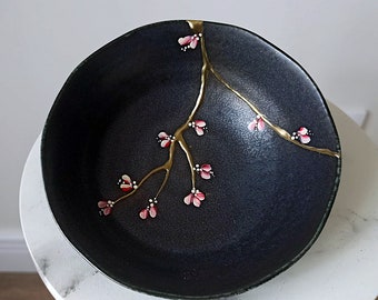 Puede incluir: Un cuenco de cerámica negro con un diseño floral dorado y rosa. El cuenco tiene una grieta en el esmalte, que se destaca con pintura dorada.