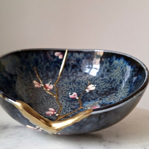 Peut inclure: Un petit bol en c&eacute;ramique de forme ovale avec un gla&ccedil;age bleu fonc&eacute; et une finition craquel&eacute;e dor&eacute;e. Le bol pr&eacute;sente une d&eacute;licate branche dor&eacute;e avec des fleurs roses peintes &agrave; l'int&eacute;rieur.