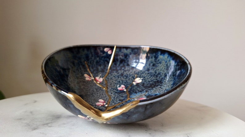 Peut inclure: Un petit bol en c&eacute;ramique de forme ovale avec un gla&ccedil;age bleu fonc&eacute; et une finition craquel&eacute;e dor&eacute;e. Le bol pr&eacute;sente une d&eacute;licate branche dor&eacute;e avec des fleurs roses peintes &agrave; l'int&eacute;rieur.
