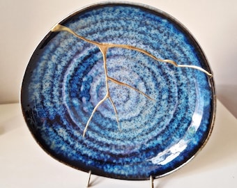 Puede incluir: Un plato de cerámica azul y blanco con una grieta dorada que atraviesa el centro. El plato tiene un patrón de remolino y se muestra en un soporte.