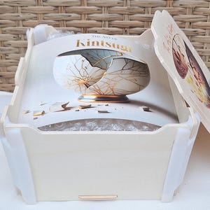 Peut inclure: Une bo&icirc;te en bois blanche avec un couvercle. Le couvercle est imprim&eacute; avec une image d'un bol en c&eacute;ramique cass&eacute; avec des accents dor&eacute;s. Le texte "The Art of Kintsugi" est imprim&eacute; sur le couvercle.