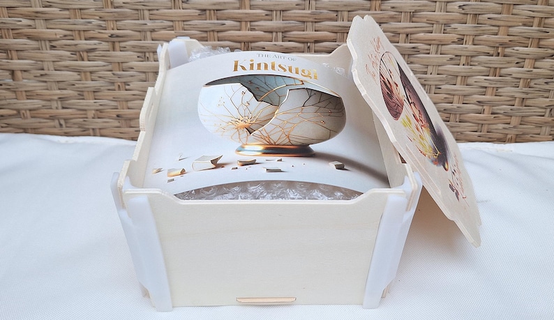 Peut inclure: Une bo&icirc;te en bois blanche avec un couvercle. Le couvercle est imprim&eacute; avec une image d'un bol en c&eacute;ramique cass&eacute; avec des accents dor&eacute;s. Le texte "The Art of Kintsugi" est imprim&eacute; sur le couvercle.