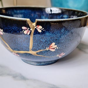May include: A blue ceramic bowl with a gold crackle glaze and pink and white floral accents. The bowl is decorated with a gold branch design.