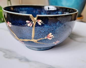 Puede incluir: Un cuenco de cerámica azul con un esmalte craquelado dorado y detalles florales rosas y blancos. El cuenco está decorado con un diseño de rama dorada.