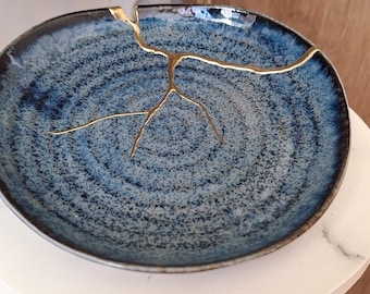 Puede incluir: Un plato redondo de cerámica azul con un patrón moteado y una reparación kintsugi dorada. El plato tiene un borde azul oscuro y se exhibe sobre una superficie blanca similar al mármol. El diámetro del plato es de aproximadamente 25 cm.