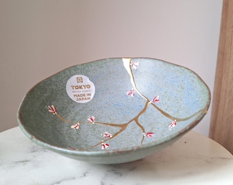 Puede incluir: Cuenco de cerámica ovalado con esmalte azul claro y verde, con reparaciones kintsugi doradas y delicadas ramas con flores rosas. El cuenco tiene una etiqueta que dice "TOKYO DESIGN STUDIO MADE IN JAPAN".