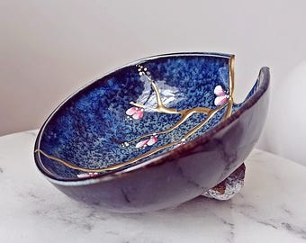 Puede incluir: Un cuenco de cerámica azul con un diseño floral dorado y rosa. El cuenco tiene una grieta en el borde y está ligeramente astillado.